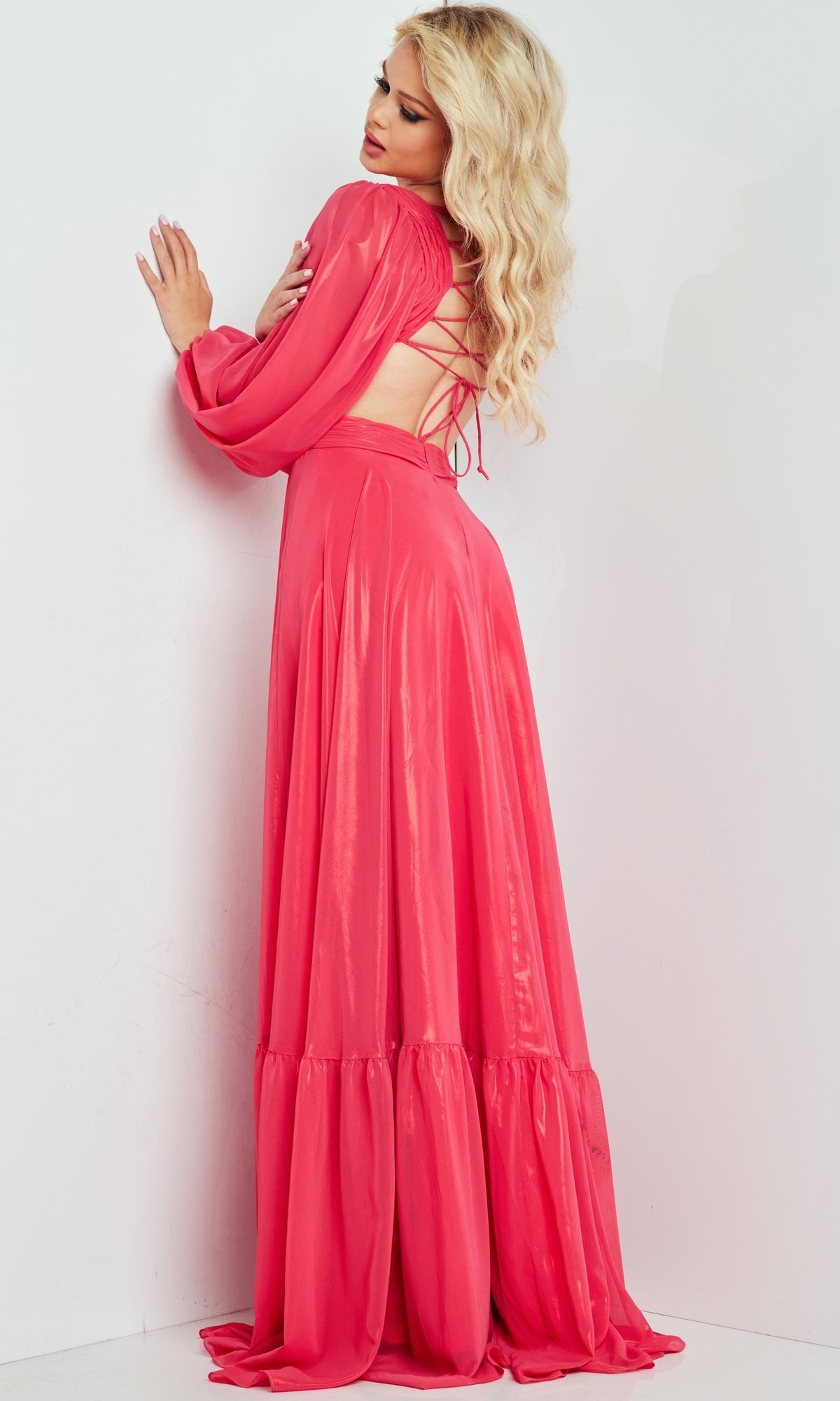 A woman with long blonde hair wears a Metallic Chiffon Long Coral Formal Dress with sleeves, featuring an open back and floor-length design, as she poses with her back to the camera against a white wall.